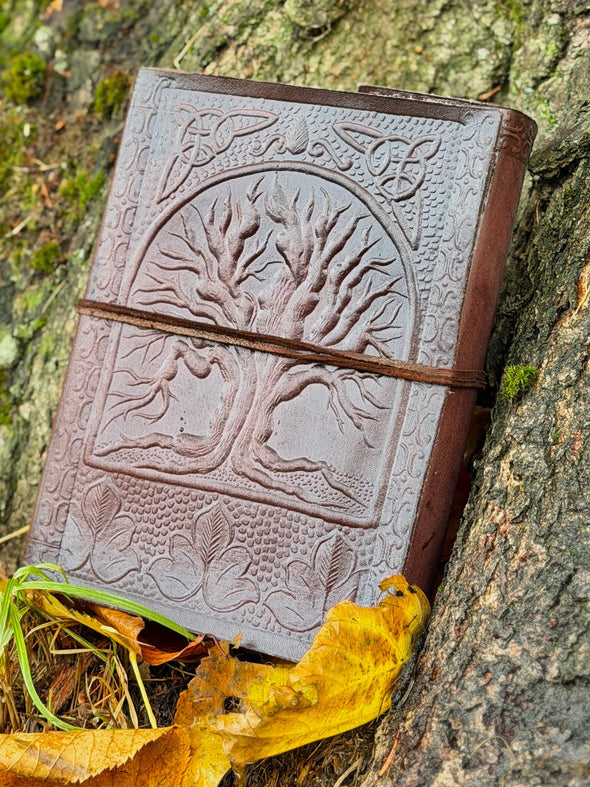 Brown leather notebook with tree design attached to a tree trunk with autumn leaves.